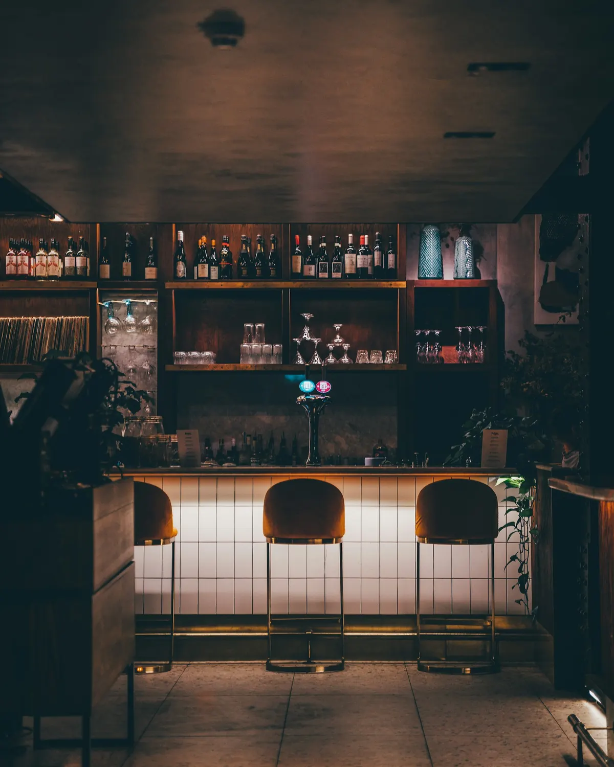 Een lege bar in de avond: drie krukken aan een verlichte toog, flessen op de planken erachter
