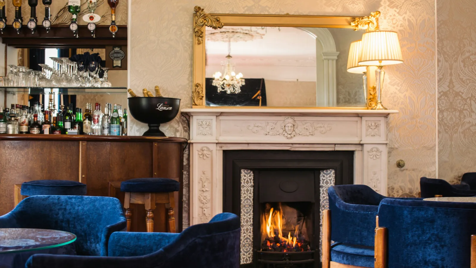 Een lounge met een brandende open haard onder een vergulde spiegel, nachtblauwe fluwelen fauteuils en een houten bar vol flessen.