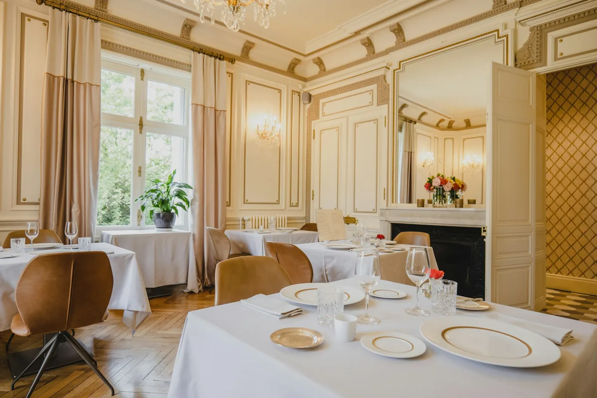 Een lichte eetzaal in een grand hotel, met witte tafellakens, porselein met gouden rand en een marmeren schouw